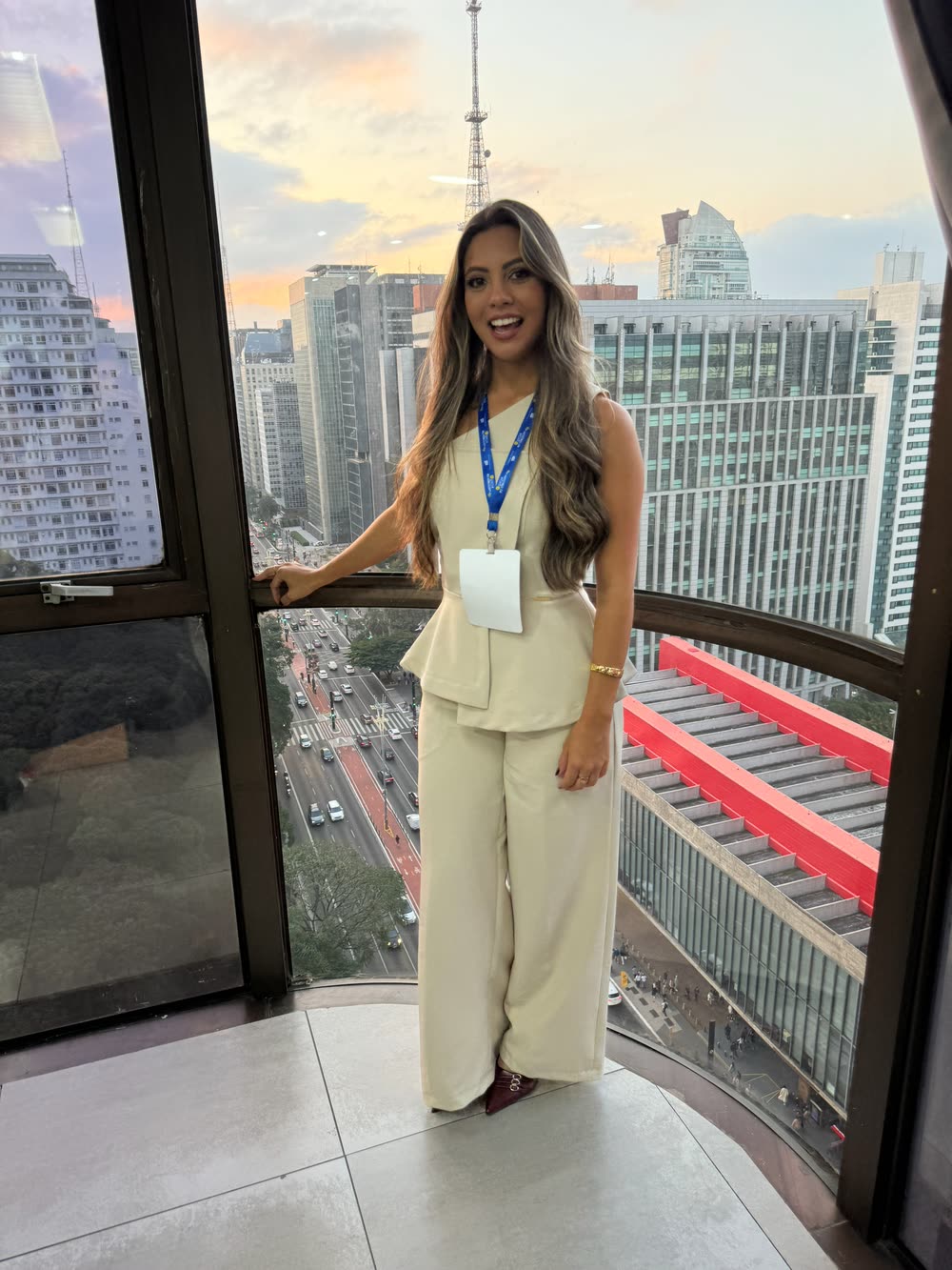 Participante de crachá sorrindo no alto de um prédio, com a Avenida Paulista e o MASP ao fundo, na edição de 2025